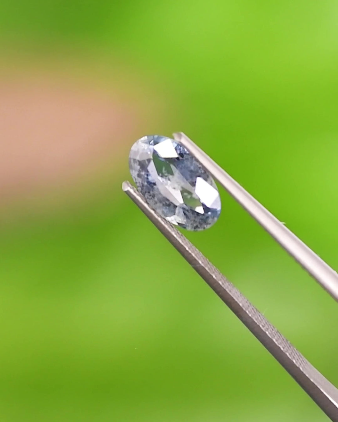 Video of lose-up of 1.24CT steely blue sapphire held in gemstone tweezers against a vivid green background, revealing elongated oval shape and untreated natural clarity from Umba River Valley, Tanzania.