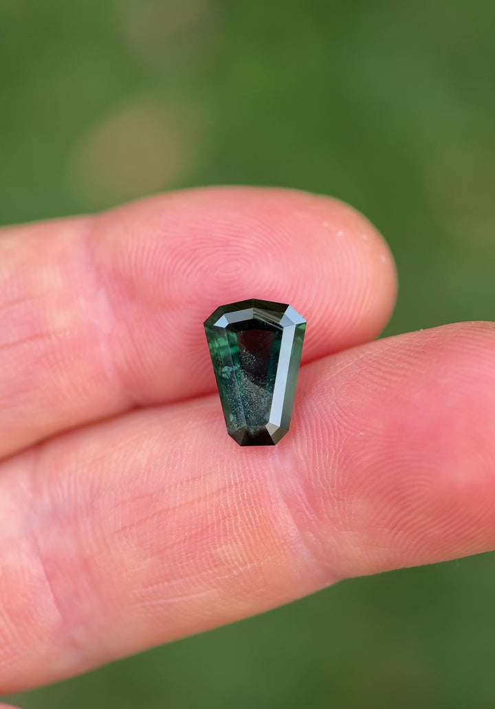 A 3.65 carat shield-cut sapphire from Garba Tula, Kenya, shown rotating in natural daylight between fingers. The deep peacock and green hues shift across its facets as grass and outdoor elements blur in the background.