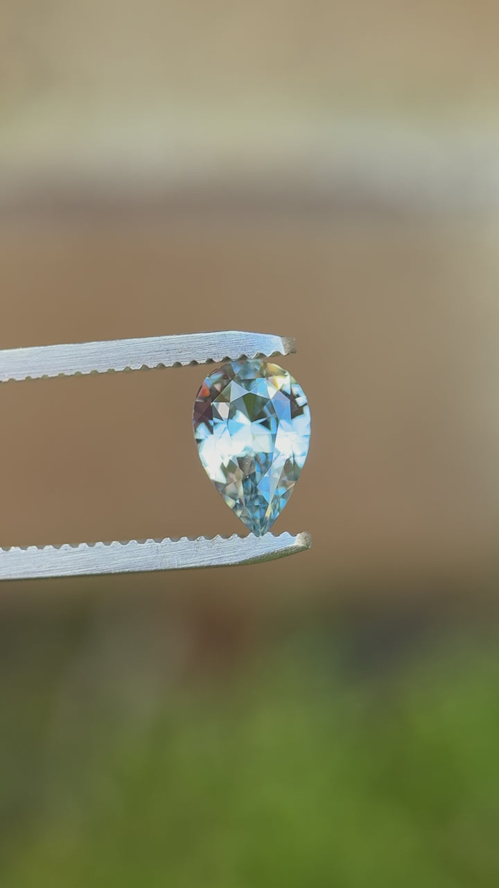 Pear-shaped Madagascar sapphire turned in tweezers under natural light, highlighting its crisp symmetry, pointed tip, and luminous blue-lavender brilliance.