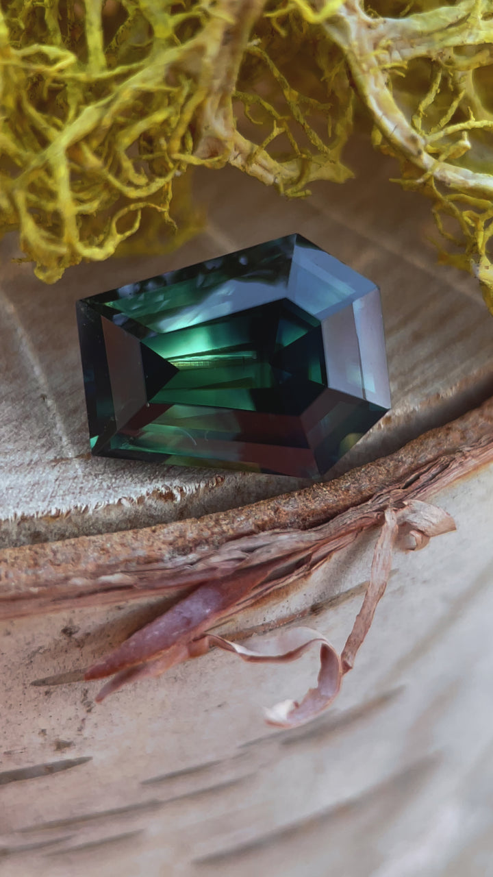 A video showcasing the dynamic brilliance of a 4.65-carat forest green sapphire in a step cut coffin shape, featuring teal and yellow hues, under natural lighting.
