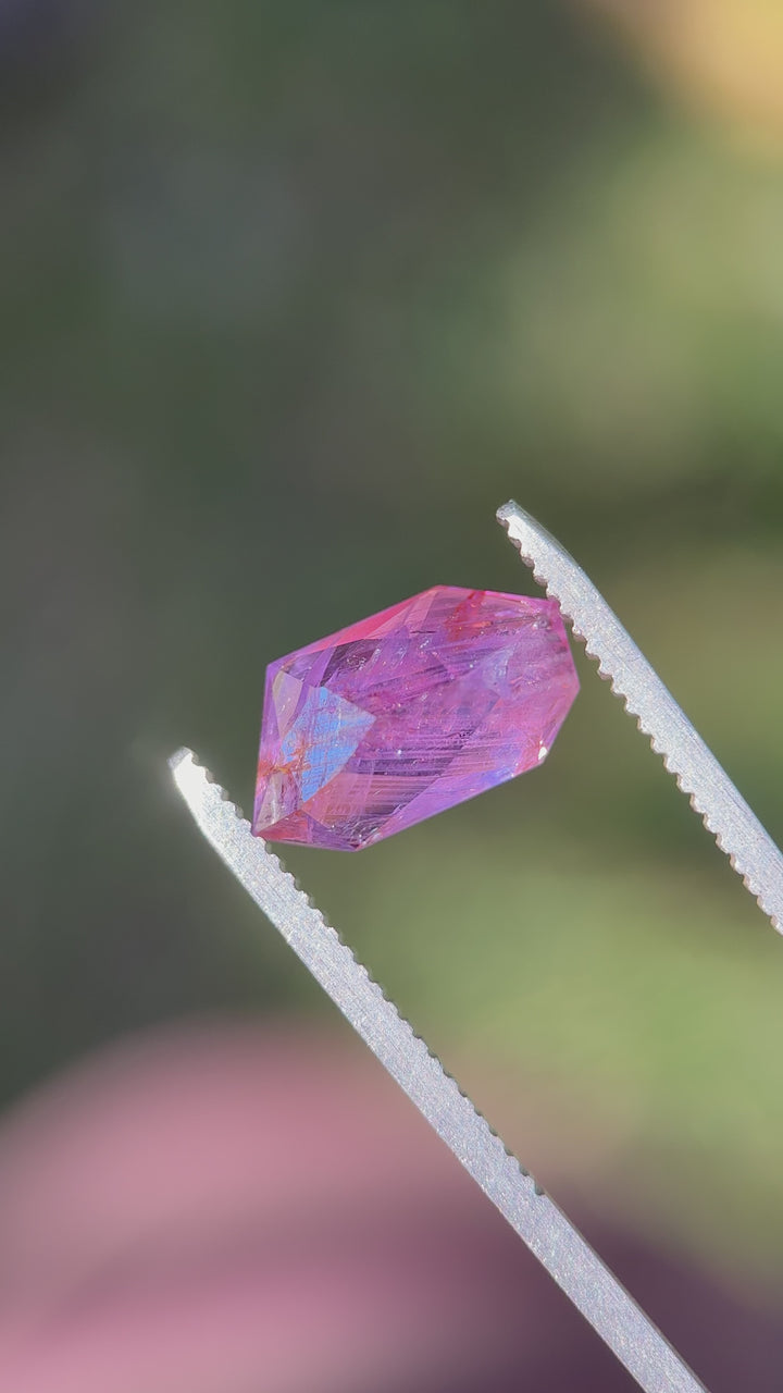 5.10ct free-form Umba sapphire turned in tweezers under natural light, showing asymmetrical facets and rich pink-to-purple color zoning.