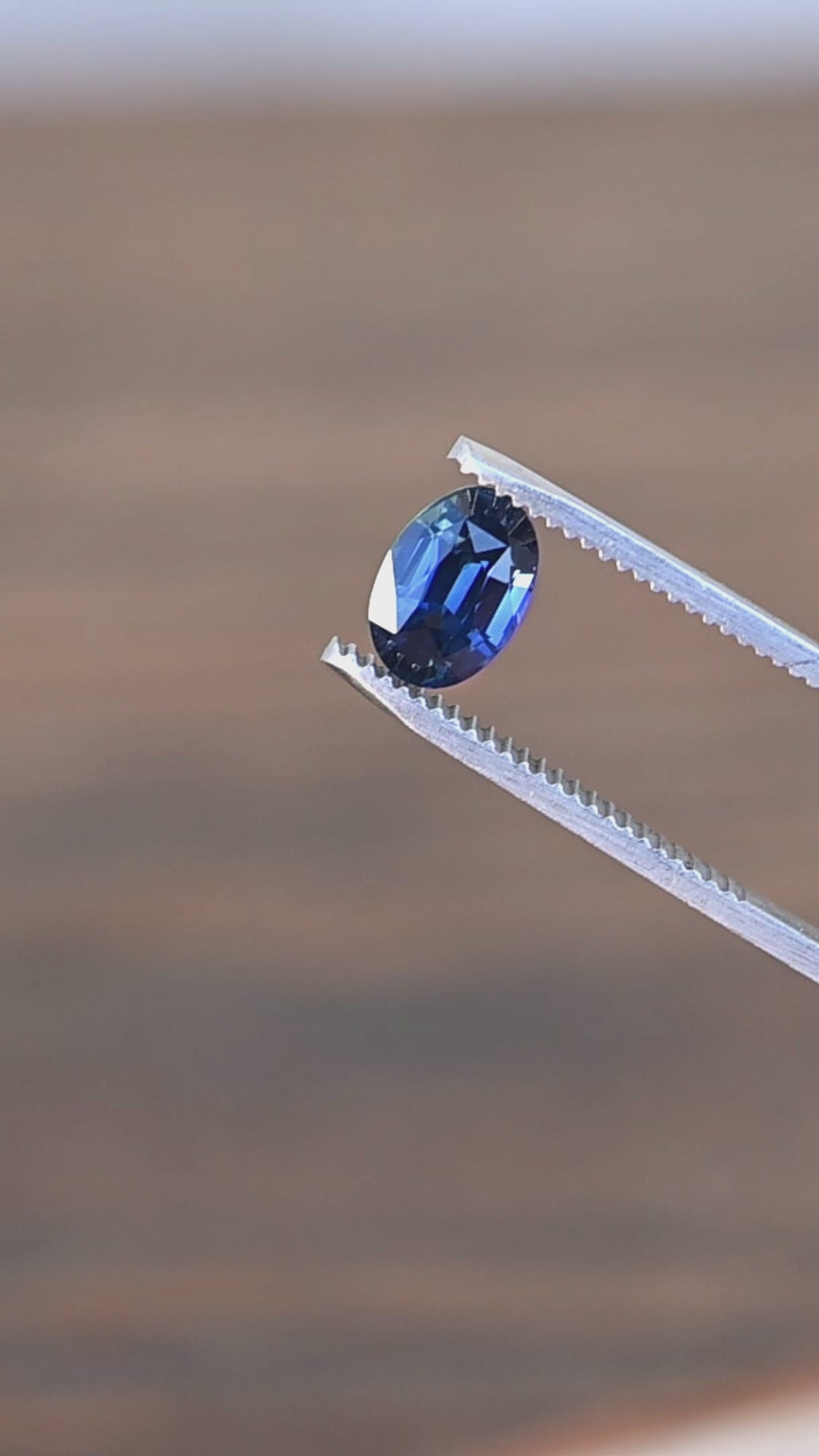 2.29ct Deep oceanic blue oval mix cut sapphire rotating in gem tweezers under natural light.