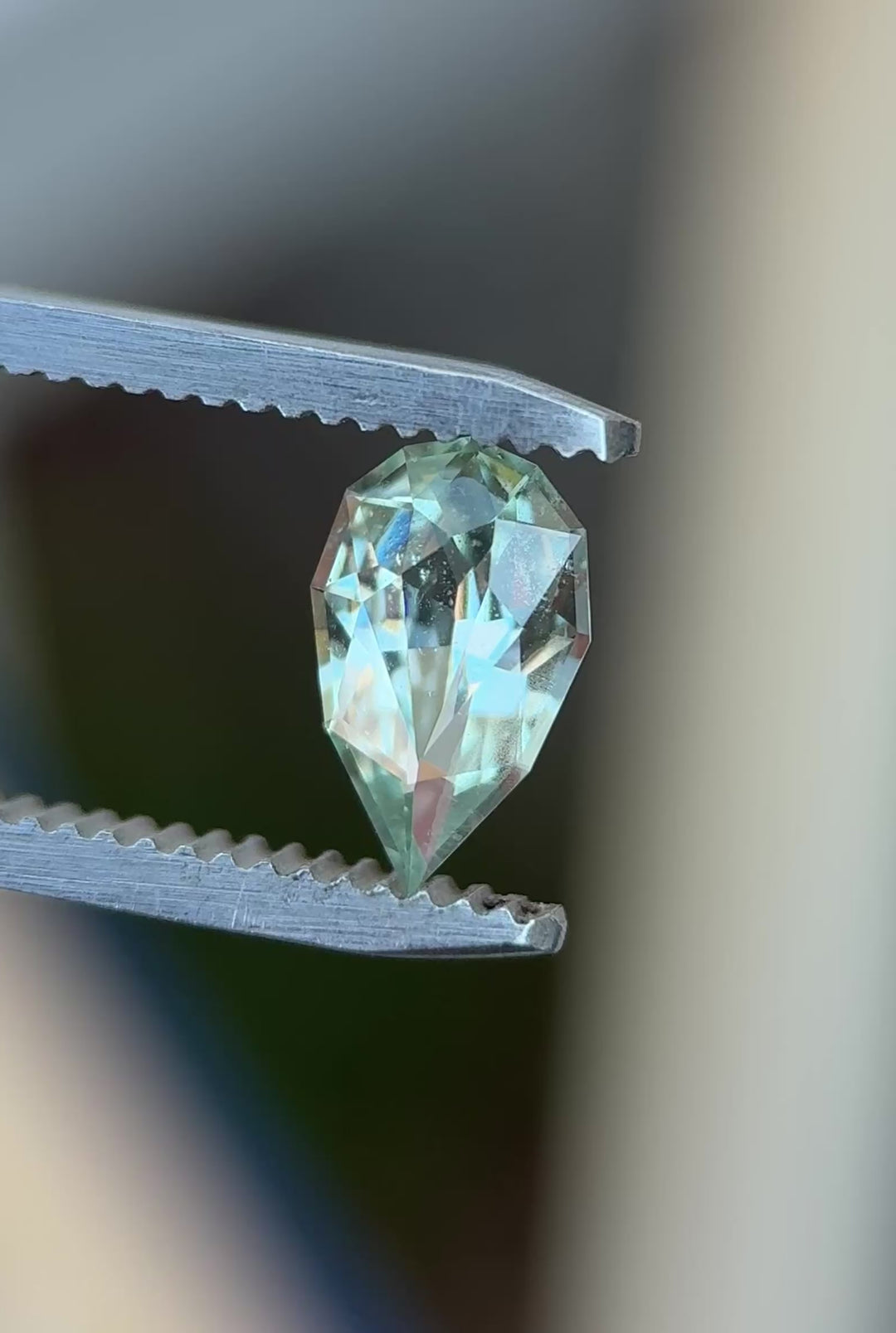 Natural Madagascar sapphire turning in tweezers, showcasing pear cut brilliance and delicate seafoam green color under changing light.