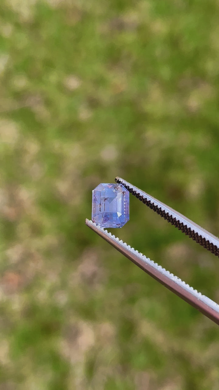 Detailed video showcasing a 1.81-carat bi-color sapphire, featuring a mix of lavender and blue hues. The video captures the gemstone's emerald cut and pristine facets as it rotates, illustrating its origin from Tanzania and emphasizing its untreated, pure form.