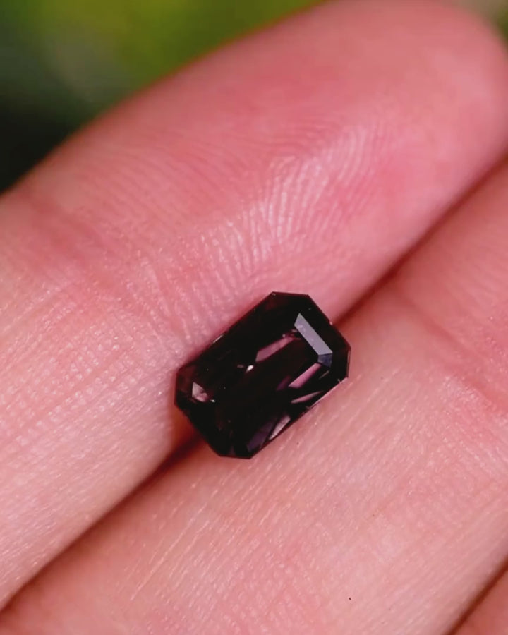 Video of 1.70CT brown violet purple sapphire displayed on fingertip, rotating to reveal flashes of purple and brown hues in its modified emerald cut, untreated gem from Umba River Valley, Tanzania.