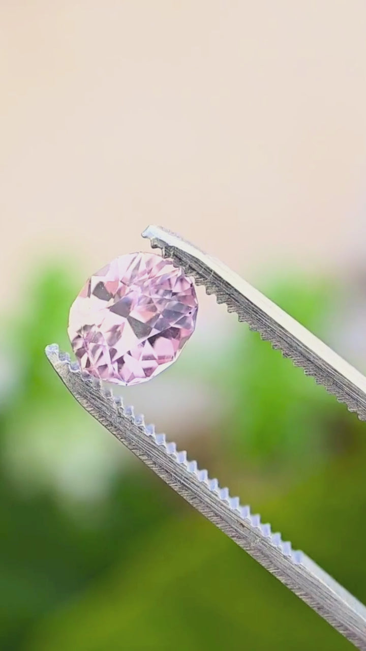 2.50 carat Sri Lankan peach pink sapphire rotating in jeweler’s tweezers, showing the stone’s custom geometric oval outline, brilliant faceting and changing blush-pink reflections in natural light.