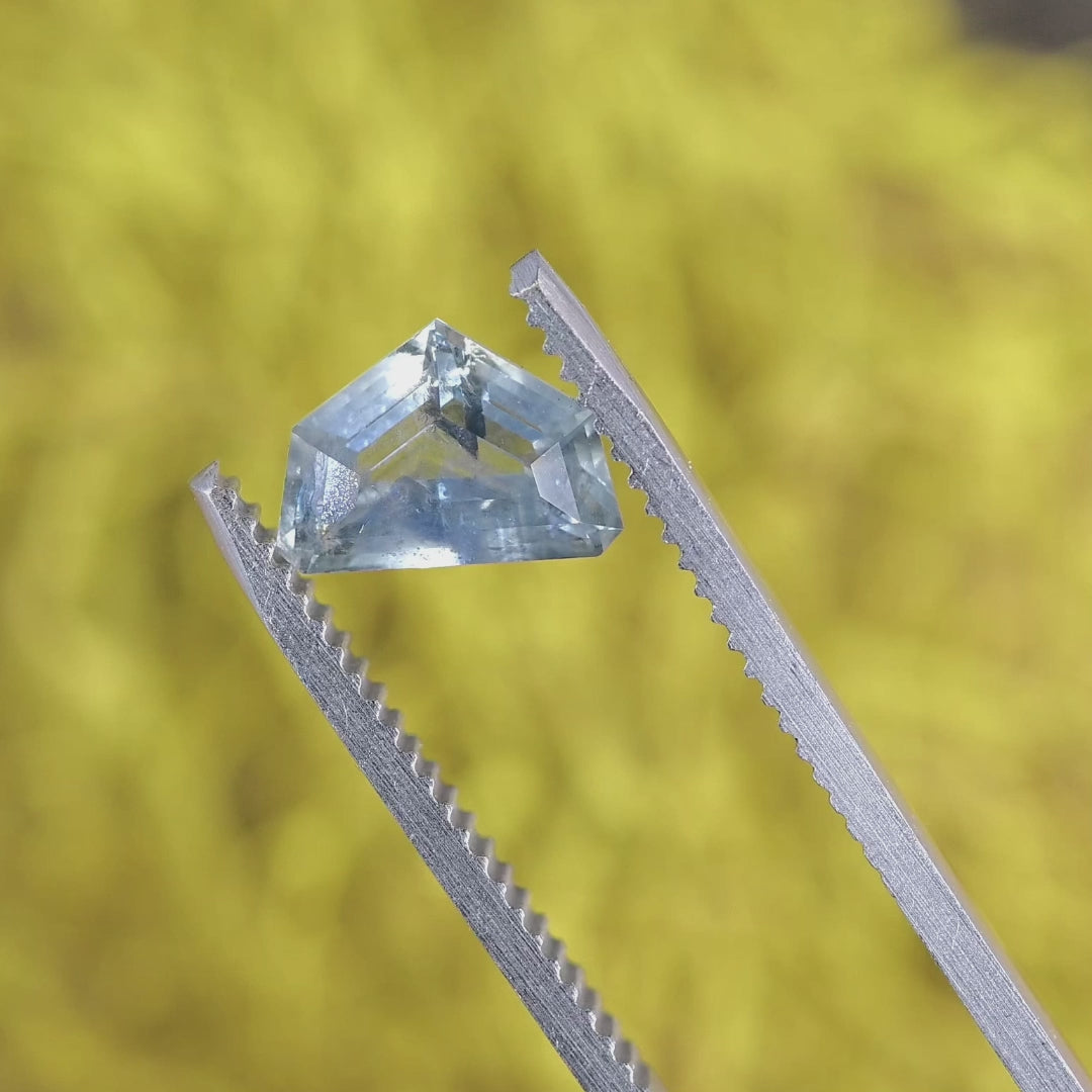 2.04ct geometric half moon sapphire in tweezers, revealing angular facet structure, transparent light blue body color, and internal inclusions visible as the stone tilts.