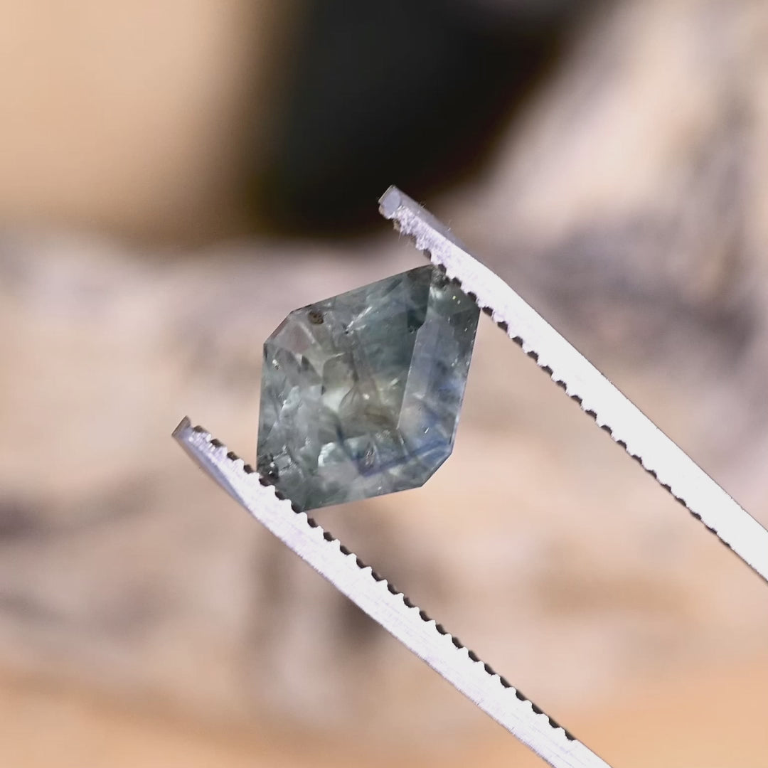 Geometric kite-shaped Montana sapphire held in metal tweezers and turned under natural light, revealing structured tablet-style faceting, layered teal-to-blue zoning, and subtle internal feathering that becomes more pronounced as the pavilion angles reflect light across the broad planes.