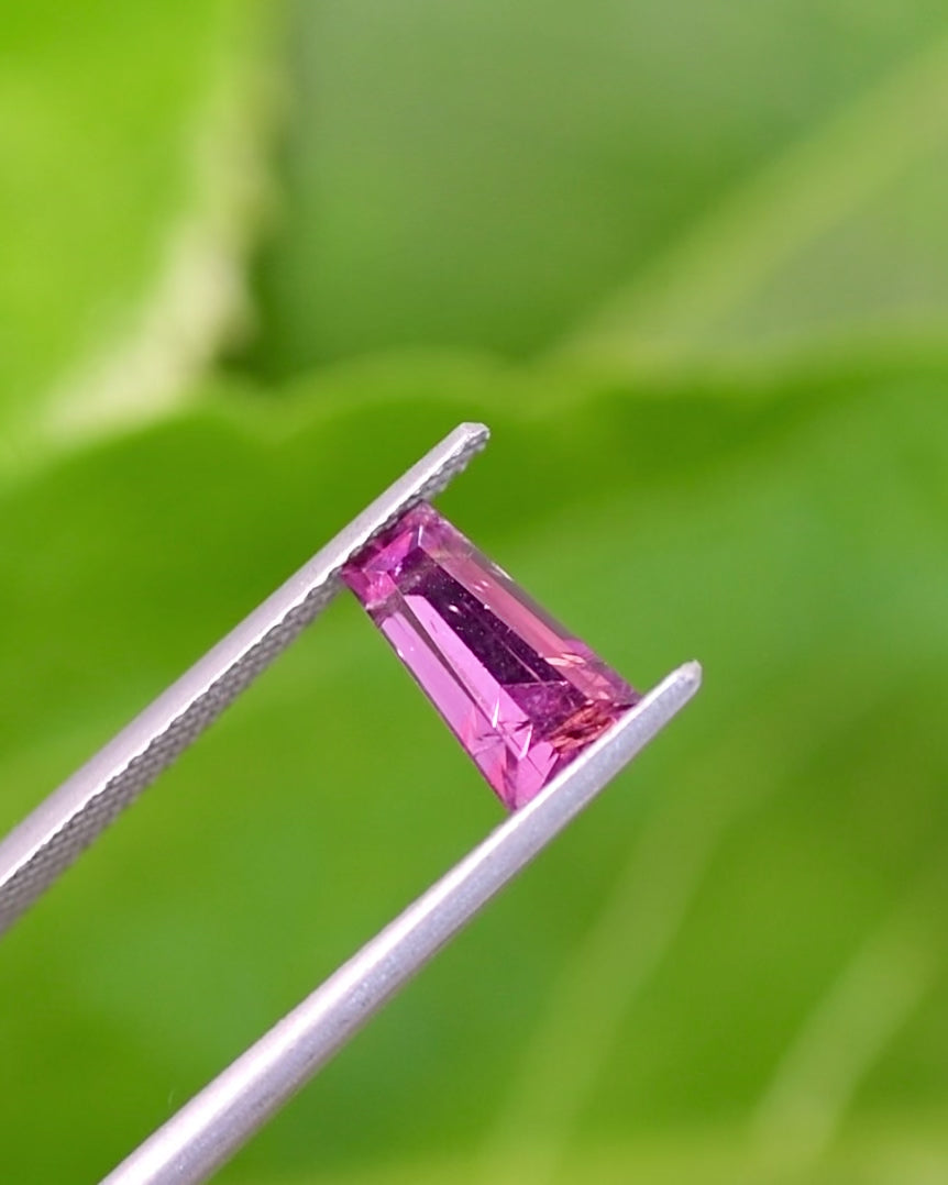 Video of 1.39CT pink and purple garnet rotating in gemstone tweezers under natural light, displaying reflections, step cut brilliance, and saturation of the tapered baguette, untreated garnet from Kenya.