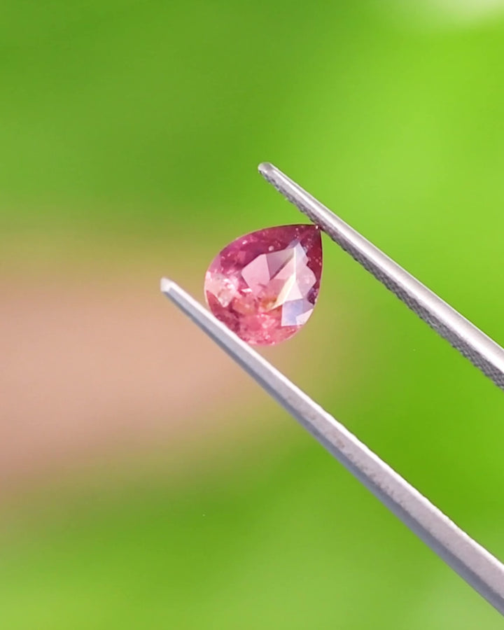 Close-up Video of 1.24CT orange pink red sapphire balanced on fingertip, showing the pear-shaped modified brilliant cut and internal inclusions under natural daylight, unheated gem from Umba River Valley, Tanzania.
