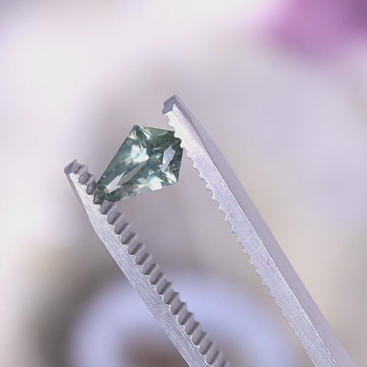 Macro video of 0.57 carat Montana sapphire kite step cut in tweezers, turning to reveal teal blue green tones, crisp facet reflections, and natural untreated sparkle