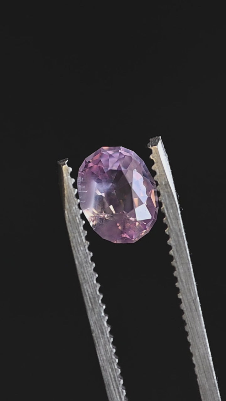 Loose oval mix cut sapphire turning in tweezers, revealing pastel pink and peach flashes with subtle violet undertones from Sri Lanka.