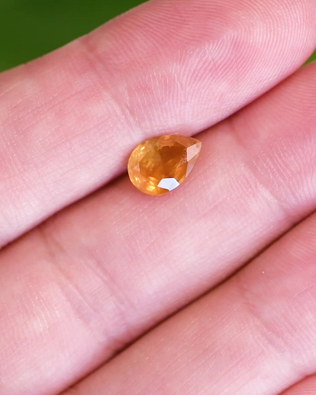 Close-up video of 1.59 carat orange sapphire resting on fingertips under natural light, capturing flashes of red-orange and yellow brilliance in the modified pear cut, untreated gem from Tanzania.