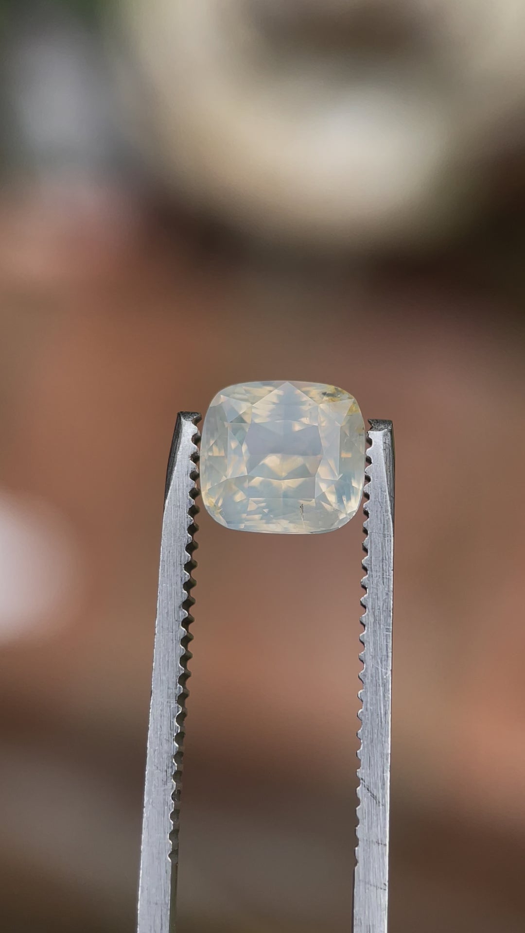 Close-up video of a 2.60-carat custom cushion-cut sapphire with a pale yellow opalescent hue, displayed in tweezers to emphasize its cut, clarity, and unique color in varied lighting.