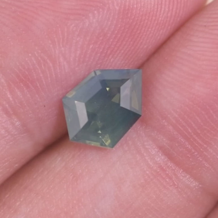 Rotating loose opalescent greenish teal blue sapphire from Diego, Madagascar on a fingertip, showing the shield step cut and shifting teal-to-green highlights in changing light.