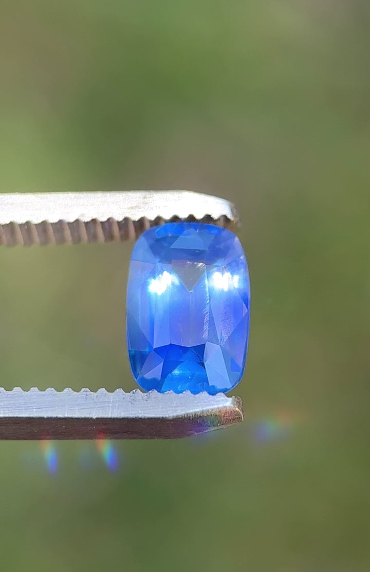 Natural Sri Lankan parti sapphire turning in tweezers, showcasing cornflower blue flashes, royal blue zoning, and elegant elongated cushion faceting.