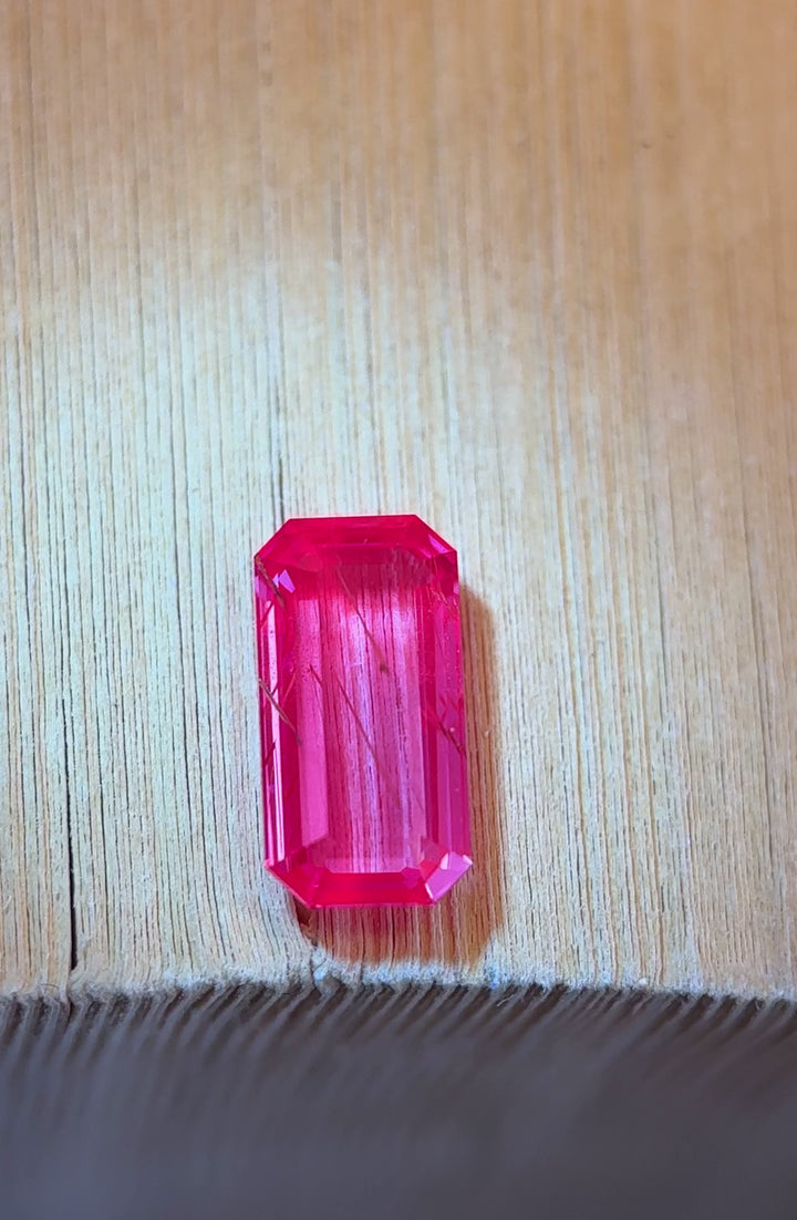 Video of a 2.01 carat rutilated purple sapphire on a wood surface, revealing elongated emerald portrait cut geometry, visible rutile needle inclusions, natural color zoning, fluorescence and untreated Madagascar origin