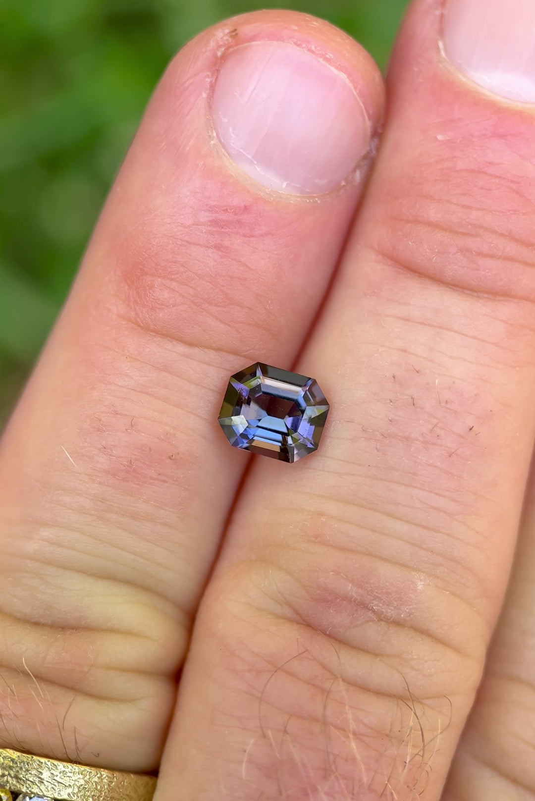 Handheld close-up video of a 1.32 carat emerald cut Tanzanite showing shifting mermaid tones of teal and purple, highlighting brilliance and scale against natural daylight.