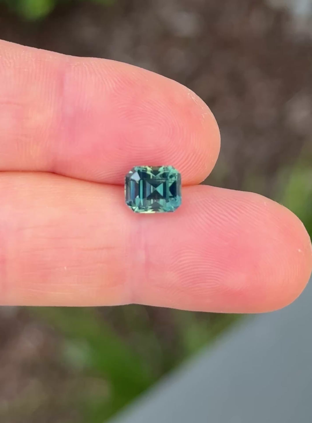 Unique teal loose sapphire with precision emerald cut faceting rotating on fingertips in natural light.