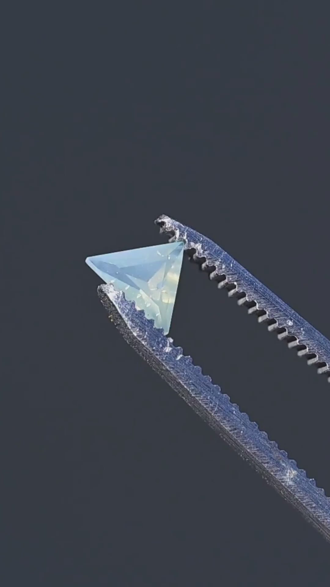 A close-up presentation of a 0.62ct untreated opalescent icy seafoam blue sapphire from Nigeria. Held in jeweler's tweezers, the gemstone features a precision custom trillion cut, pale aqua-blue coloration and softly diffused internal appearance.