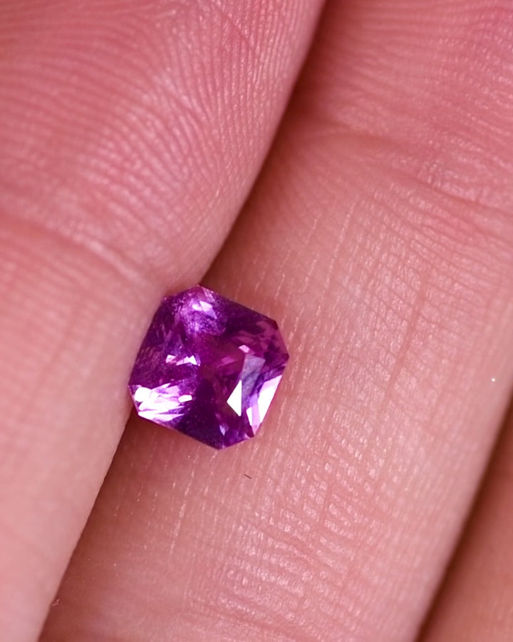 Close-up video of 1.03 carat pink-purple sapphire resting on surface, rotating under natural light to display color flashes and intricate faceting of the square scissor cut, unheated sapphire from Sakaraha, Madagascar.
