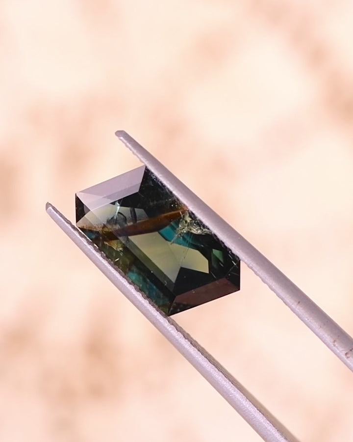 Video of 3.93CT deep peacock teal sapphire rotating in tweezers under natural sunlight, revealing flashes of blue, green, and gold through its custom corner rhombus mixed cut, unheated gemstone from Garba Tula, Kenya.