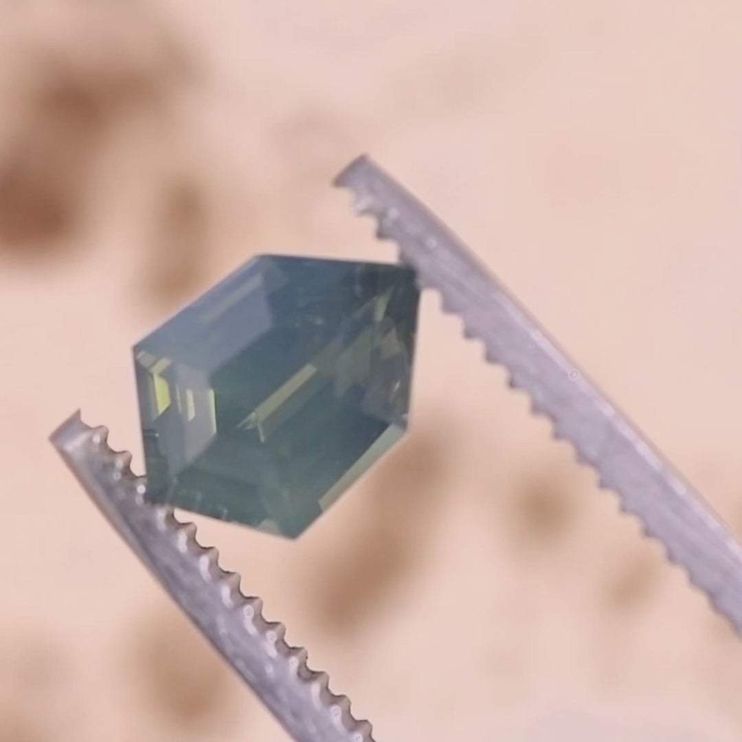 Rotating loose opalescent greenish teal blue sapphire from Diego, Madagascar in tweezers, showing the shield step cut and broad flashes as the stone turns in the light.