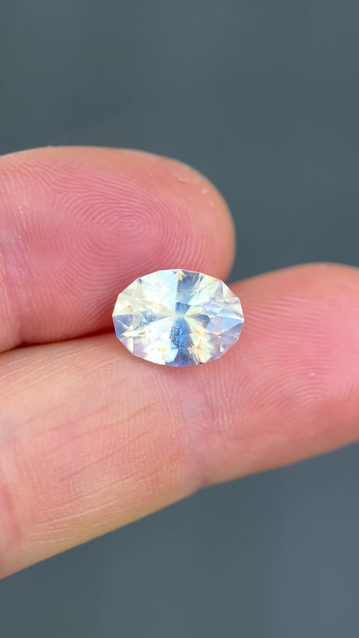Oval brilliant cut sapphire in tweezers displaying internal opalescence and subtle color movement.