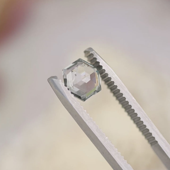 Close-up video of 1.49 carat hexagon step cut Montana sapphire in tweezers, displaying green violet teal color shift and geometric precision faceting under natural lighting.