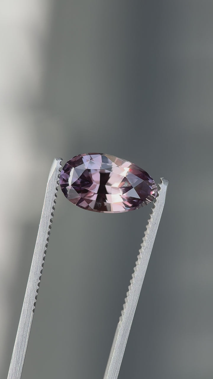 Loose heated sapphire held in tweezers showing the modified brilliant pear cut and shifting purplish red to peach champagne color.