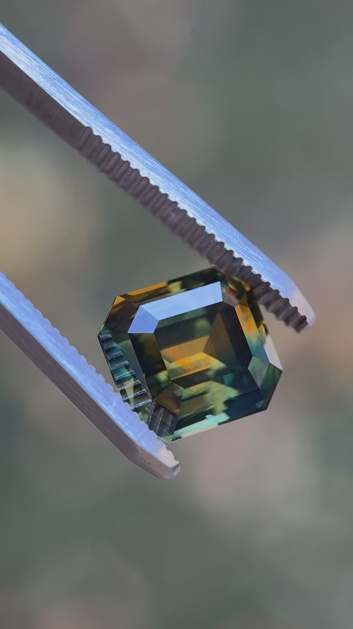 Macro video of a 9.14 carat Nigerian parti sapphire emerald cut, showing sharp facet reflections and distinct orange, yellow, blue, and green color zoning as the stone is viewed from multiple angles