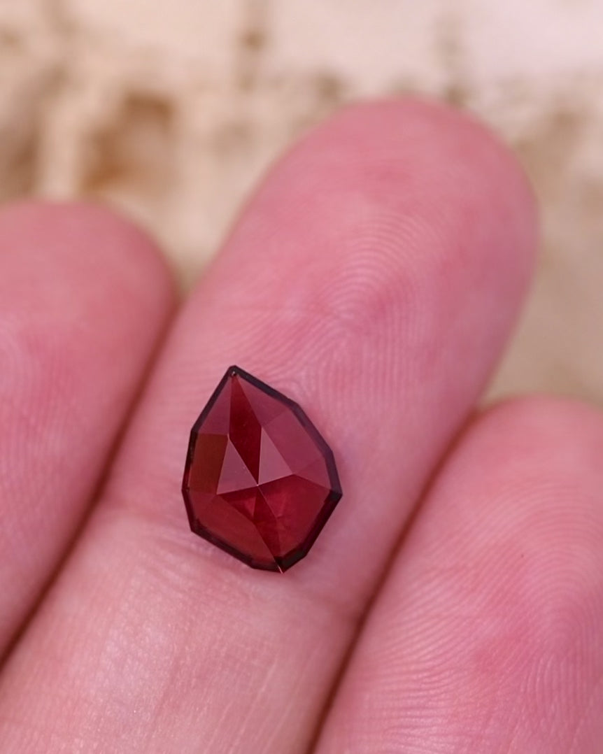 Macro video of 1.66 carat red orange garnet resting on a finger, highlighting reflections, precision facets, and light movement within the custom geometric rose cut shield, unheated garnet from Kenya.