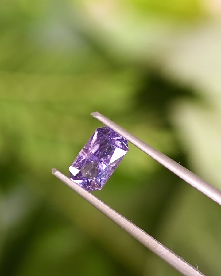Video of 0.94CT gray purple sapphire held in tweezers, rotating under natural light to reveal radiant cut facets and shifting gray-to-purple tones, unheated sapphire from Umba River Valley, Tanzania.