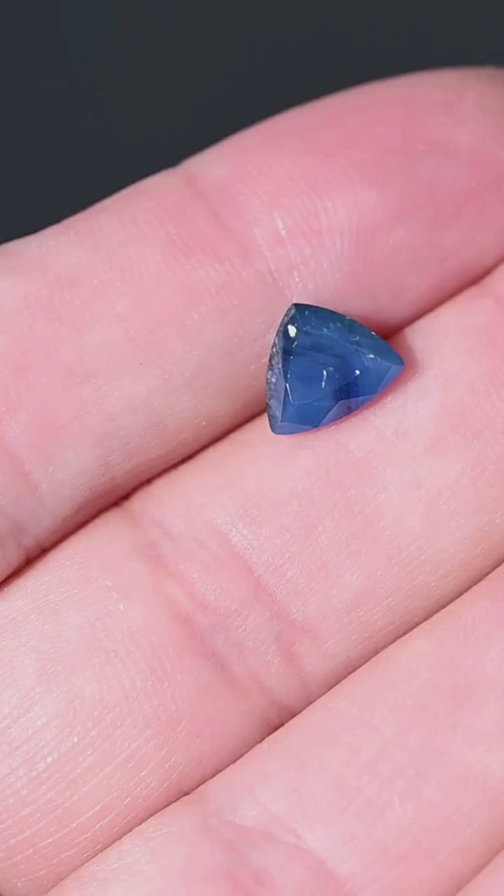 Discover a natural 1.91 carat opalescent royal blue sapphire from Gombe, Nigeria. This untreated gemstone features a custom trillion cut, distinctive internal texture and rich blue coloration. Shown against the hand for scale, it offers an unusual choice for bespoke engagement rings and custom jewelry.