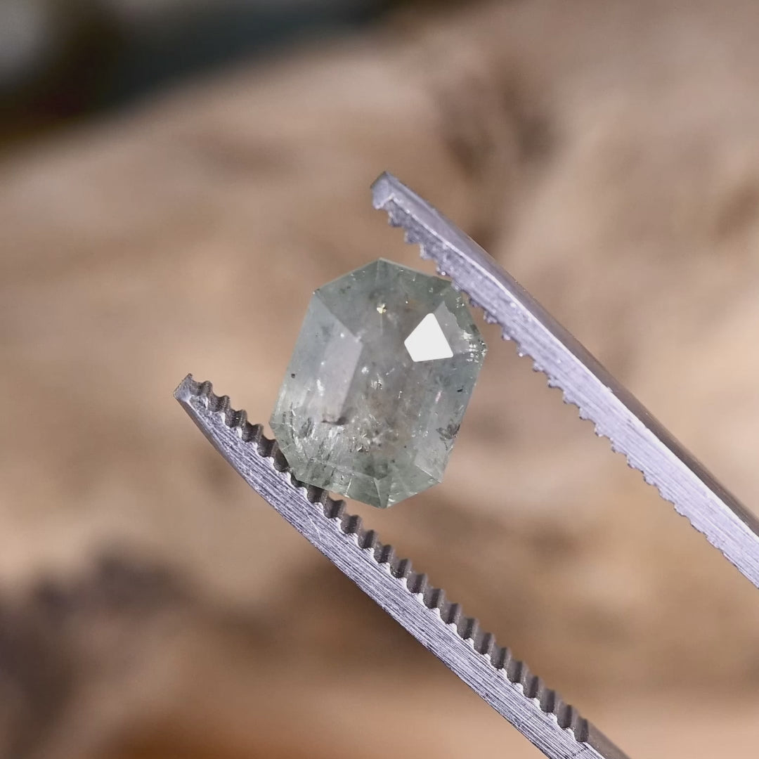 2.62ct geometric portrait cut sapphire in tweezers, showing angular facet reflections, transparent seafoam body color, and visible internal character under natural lighting.