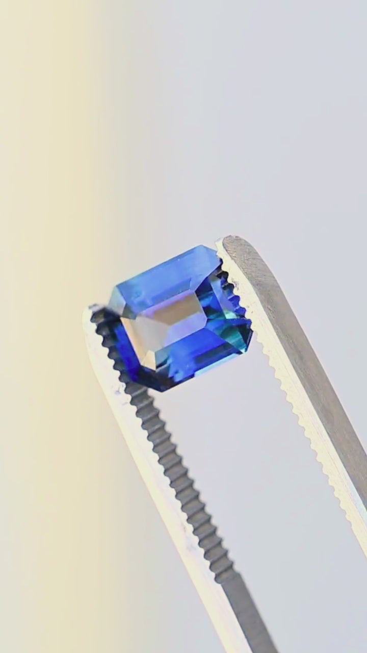 Deep royal blue Madagascar sapphire rotating in jeweler’s tweezers, showing the movement of light through its precision step facets, clipped corners and slightly teal blue color.