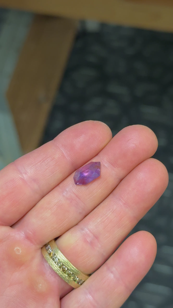 Unheated pink and purple Umba sapphire rotated in hand, revealing shifting magenta and violet tones with visible internal silk and free-form facet reflections.