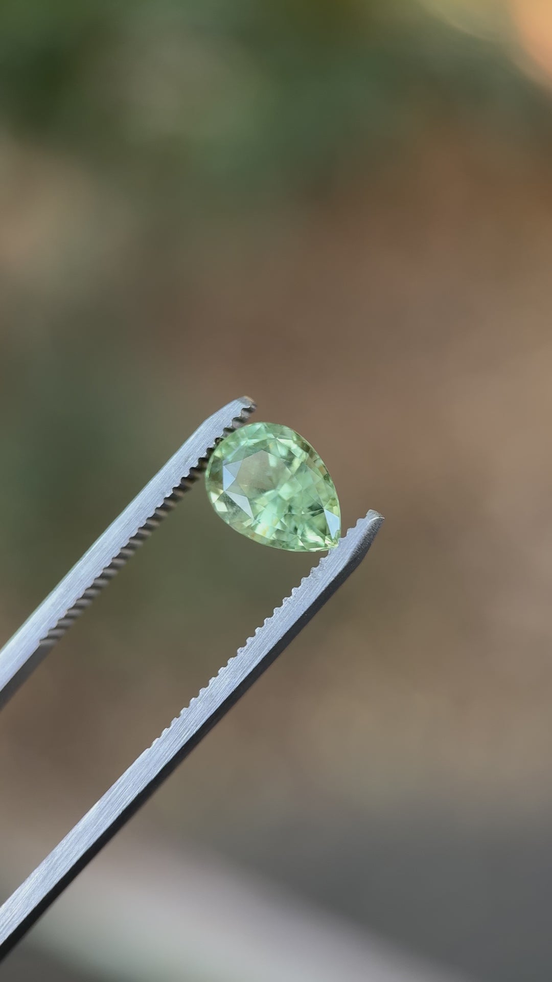 Pear-cut sapphire in earthy green and peach hues filmed in tweezers outdoors, highlighting light reflection and gemstone clarity.