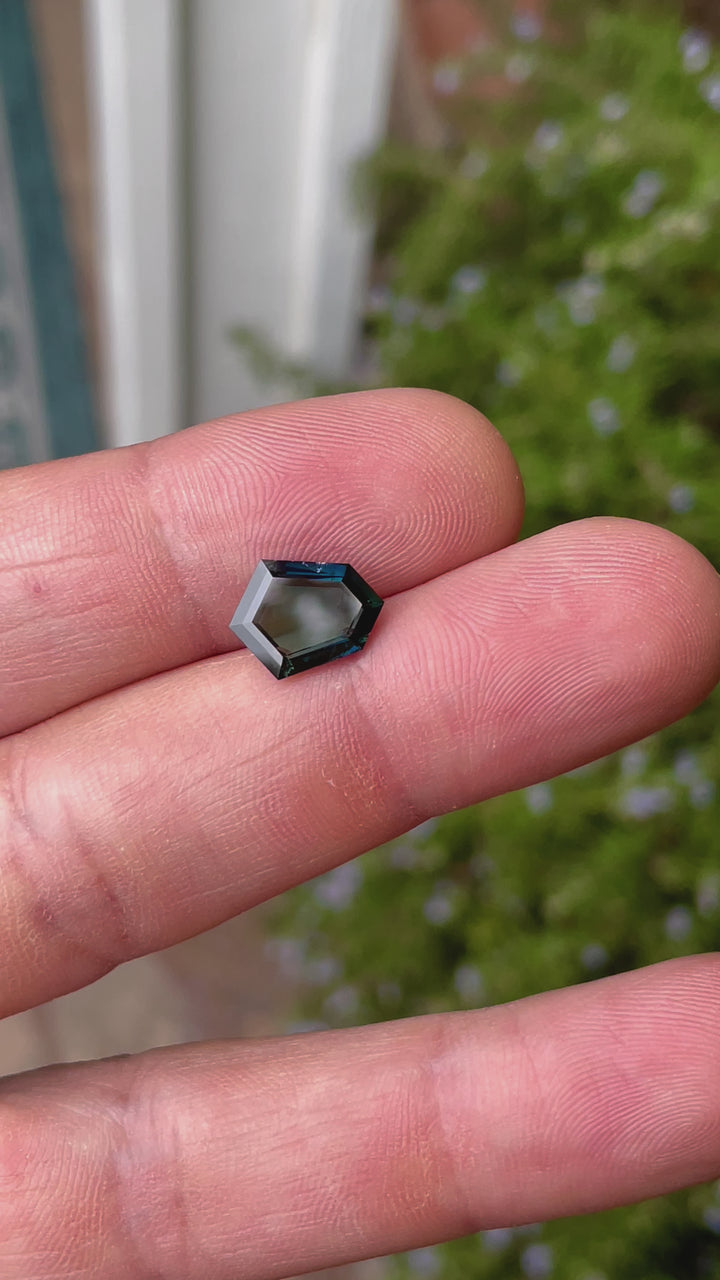 A 3.77-carat sapphire with a custom shield portrait cut is displayed under sunlight, highlighting its deep peacock, teal, blue, and green colors. The untreated gemstone, sourced from Garba Tula, Kenya, measures 12.54mm by 9.29mm by 3.02mm, and its clean facets reflect light beautifully.