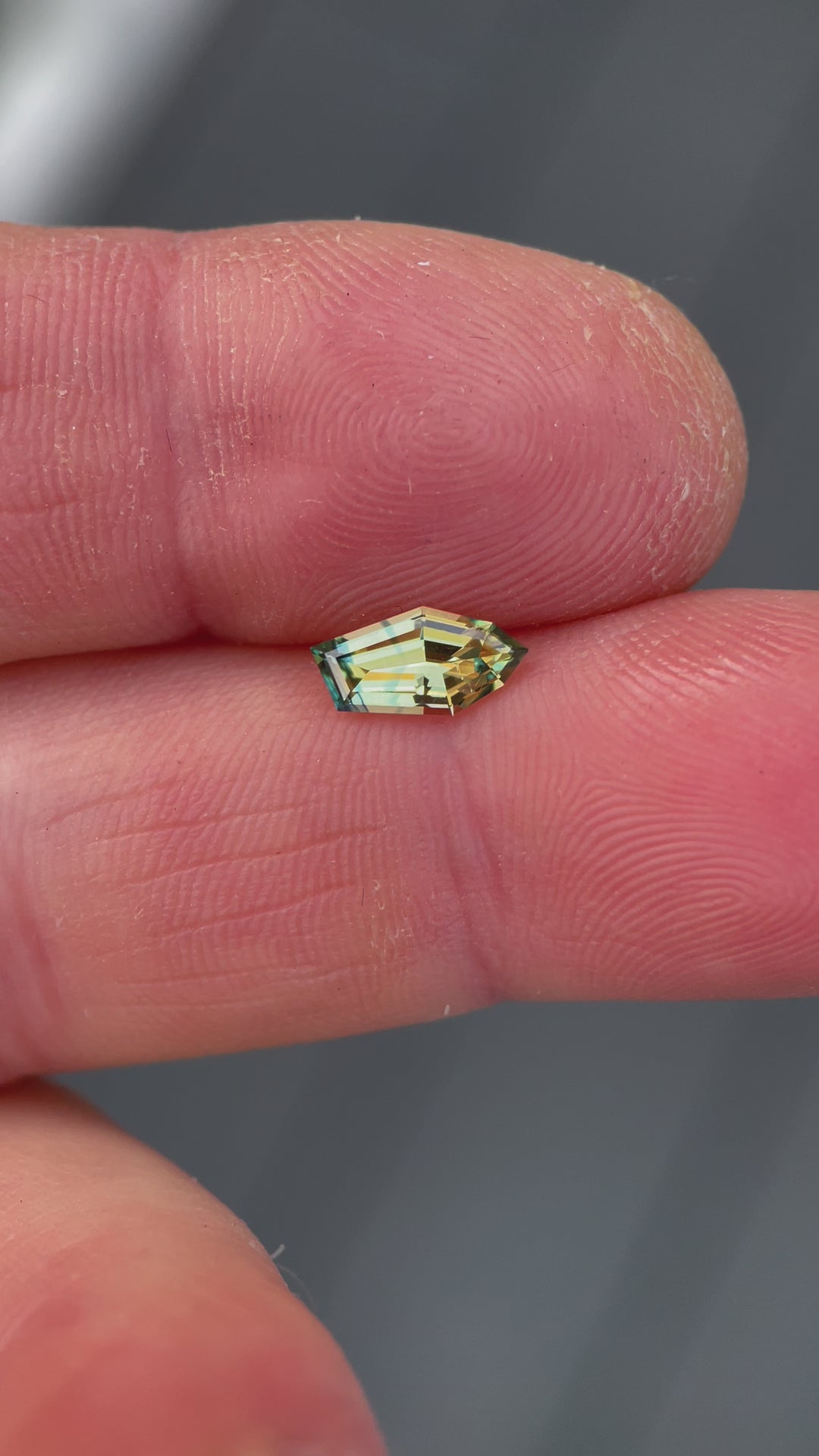 Dynamic presentation of a 0.80-carat yellow-blue sapphire, emphasizing its unique freeform geometric shape and brilliant color blend. The video captures the gemstone in natural lighting, showcasing its clarity and the distinct colors that make it a valuable piece from Nigeria.