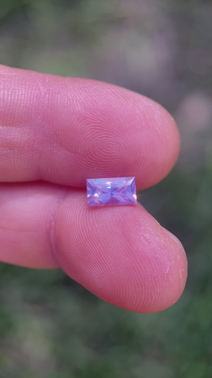 Rotating video of a baguette-cut 1.53ct pink and purple sapphire in gem tweezers, showing light refraction and color transition in natural daylight.
