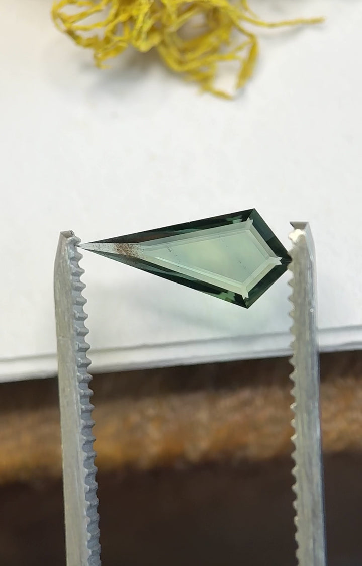 Kite portrait cut sapphire rotating in tweezers highlighting sharp edges, depth, and teal blue green tones.