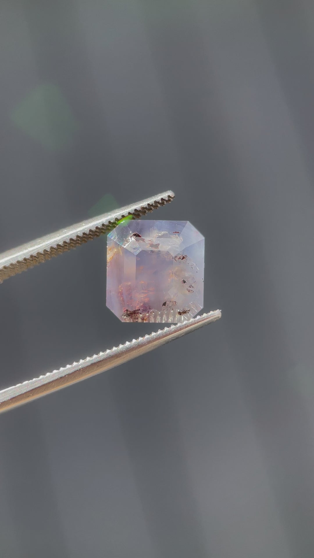 A close-up video of a 5.92-carat emerald cut sapphire, showcasing its gray, purple, and brown hues with intricate inclusions under natural light.