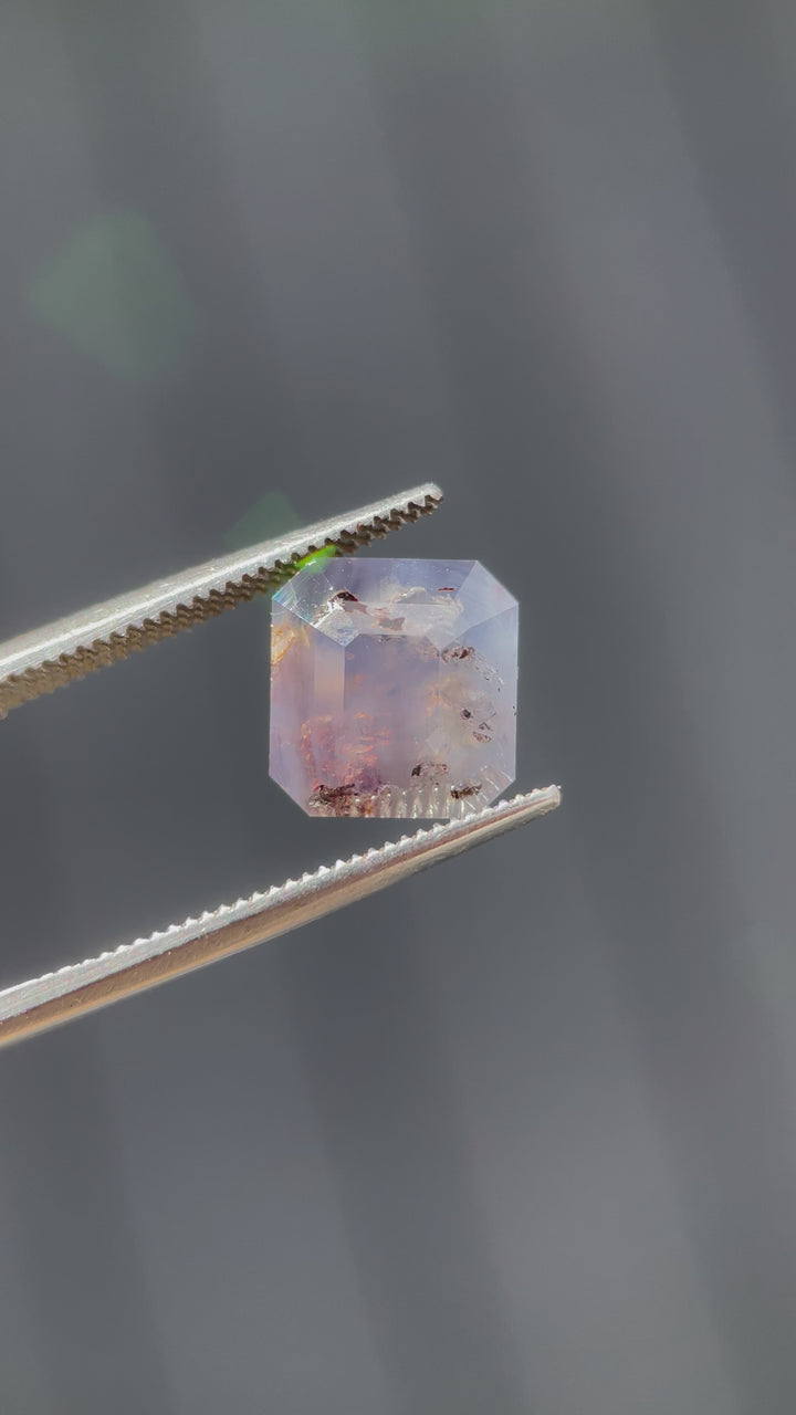 A close-up video of a 5.92-carat emerald cut sapphire, showcasing its gray, purple, and brown hues with intricate inclusions under natural light.