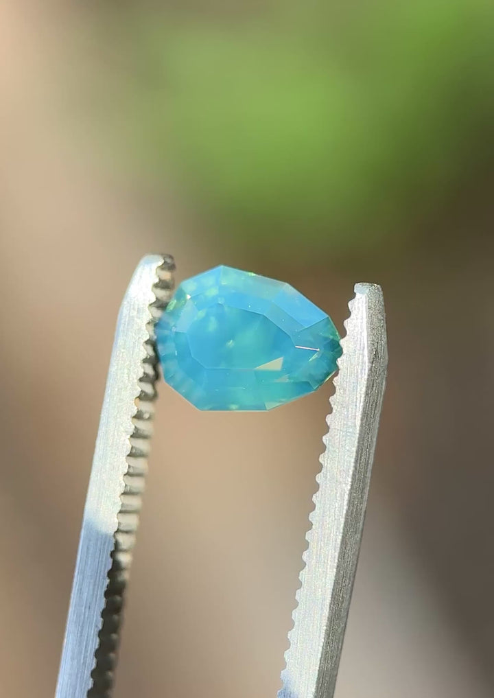 Natural Madagascar sapphire turning in tweezers, showcasing precision shield faceting and subtle teal-to-blue color movement under changing light.