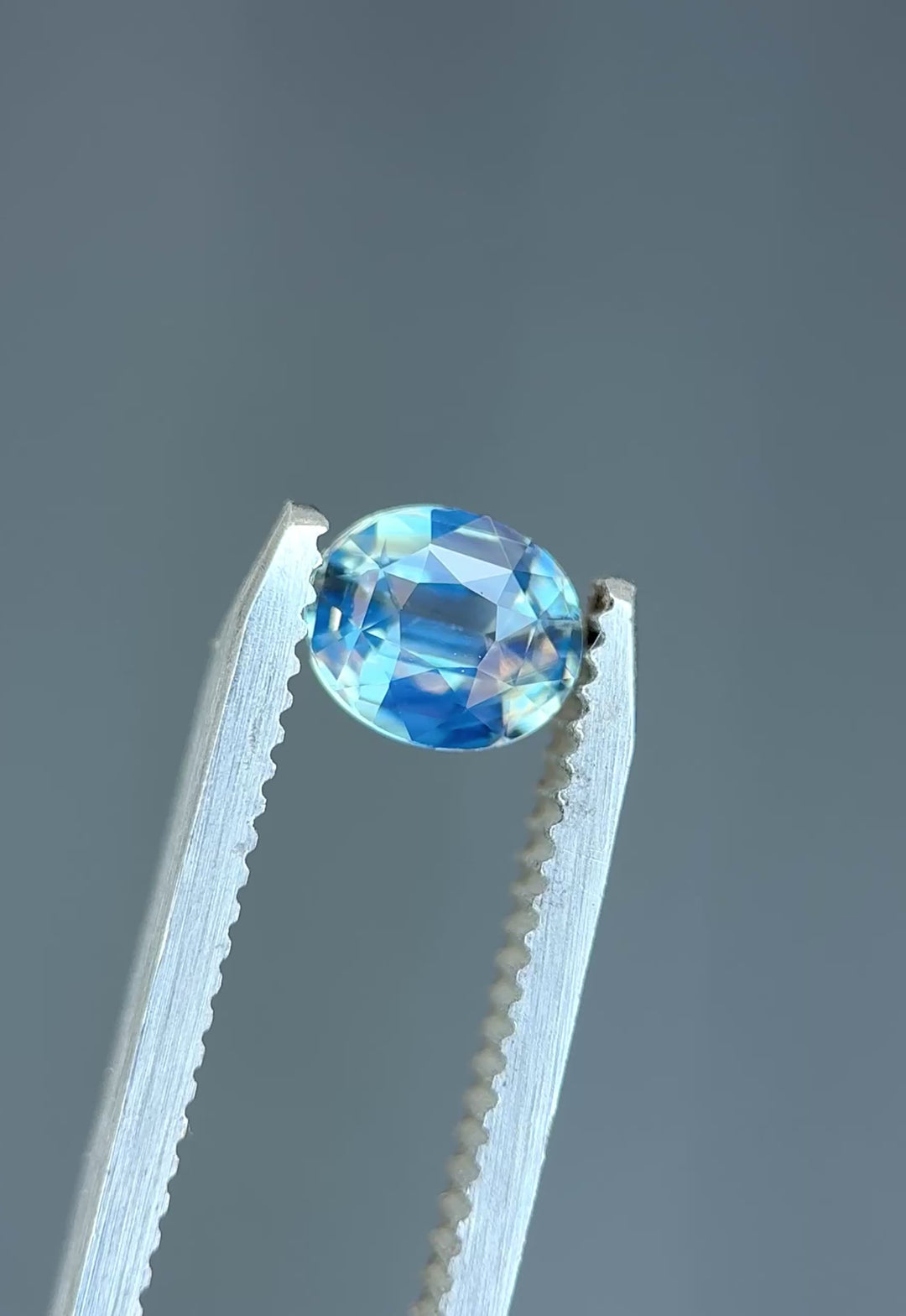 Natural Madagascar sapphire turning in tweezers, showcasing powder blue color, opalescent glow, and crisp oval symmetry.