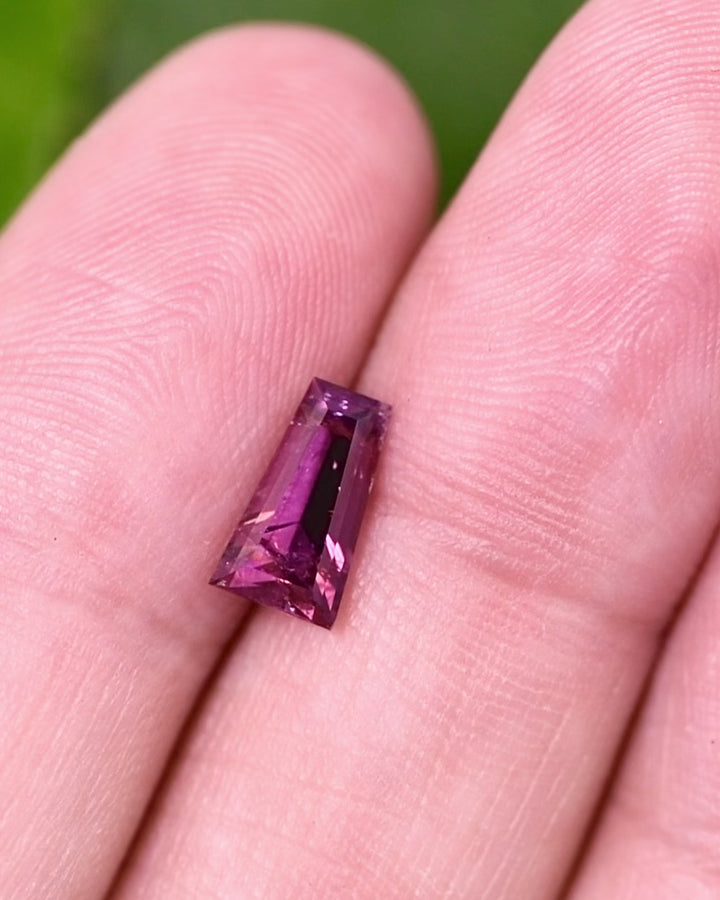 Macro video of 1.39 carat pink purple garnet resting in fingers, capturing light play, internal clarity, and detailed faceting of the step cut tapered baguette, unheated garnet from Kenya.