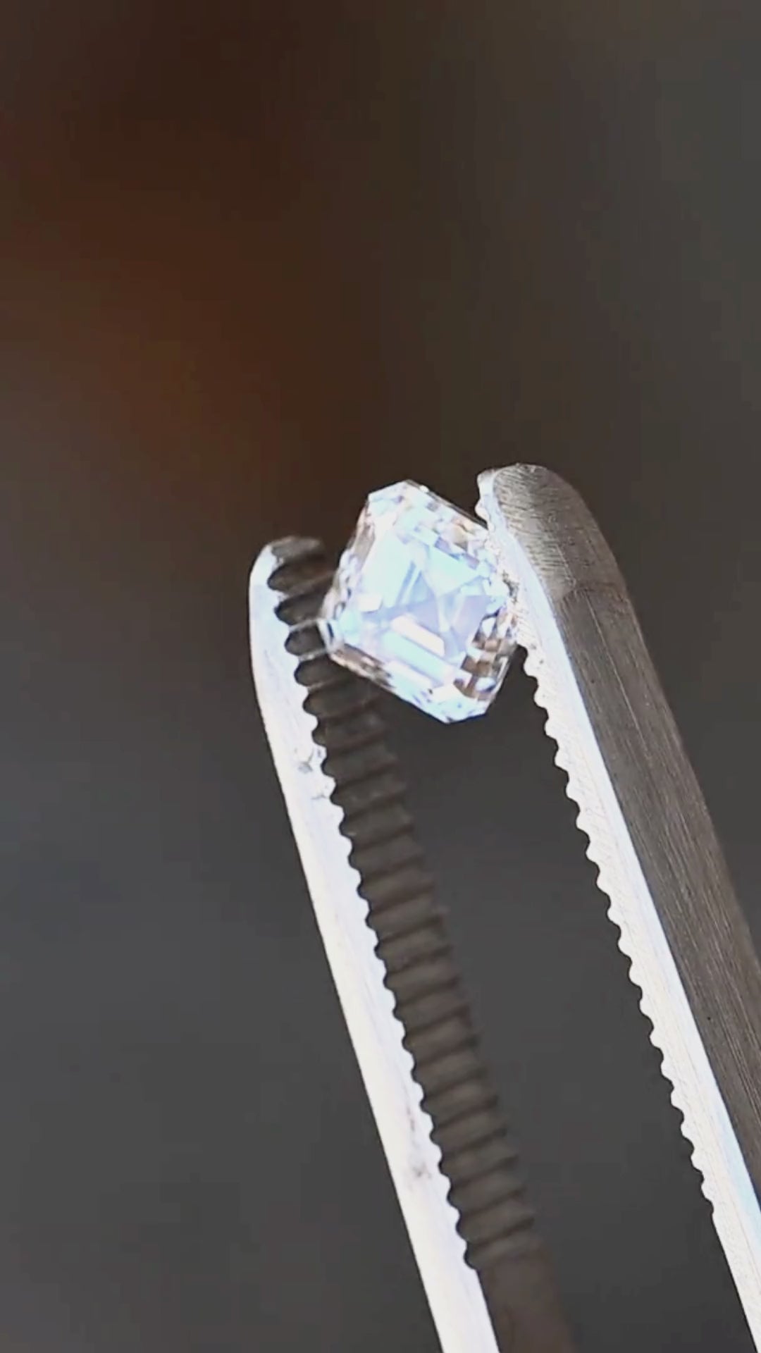1.02 carat M VVS1 Asscher cut diamond rotated in jeweler’s tweezers, showing its clipped corners, deep concentric steps and changing hall-of-mirrors reflections.