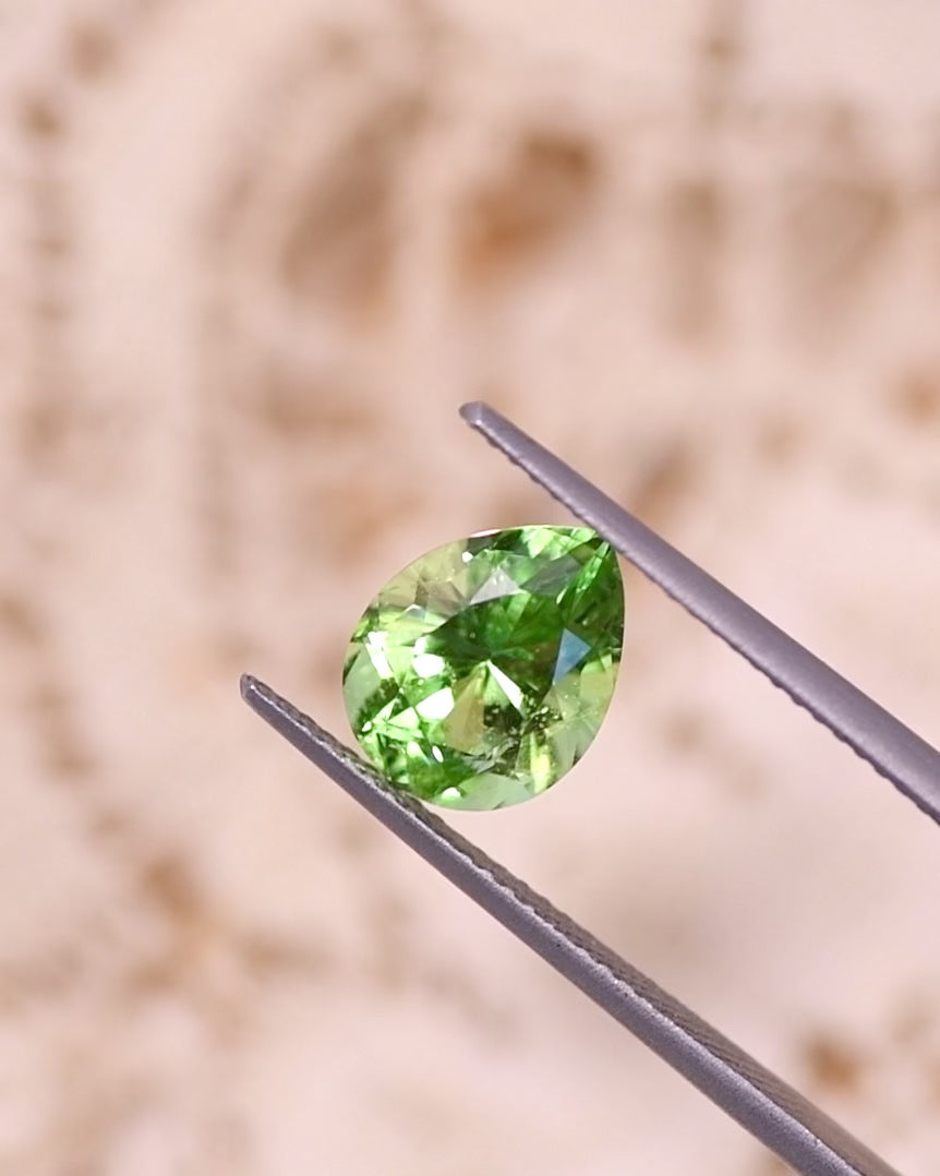 Video of 2.85CT electric lime green Tsavorite garnet rotating in gemstone tweezers under natural daylight, showcasing vivid brilliance, refraction, and color intensity of the modified brilliant pear cut, untreated Tsavorite from Kenya.