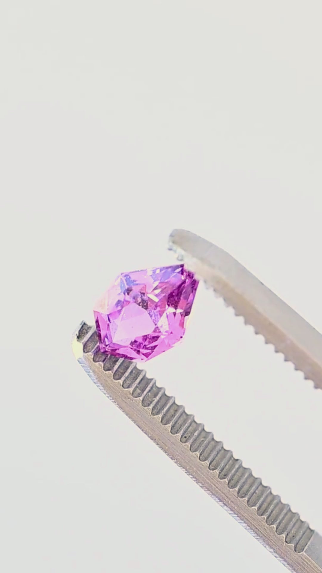 Vibrant pink-purple Madagascar sapphire rotating in jeweler’s tweezers, showing the sharp pointed outline, geometric faceting and changing flashes of saturated pink and violet.